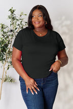 Woman wearing a black short sleeve t-shirt and blue jeans, standing indoors by a plant
