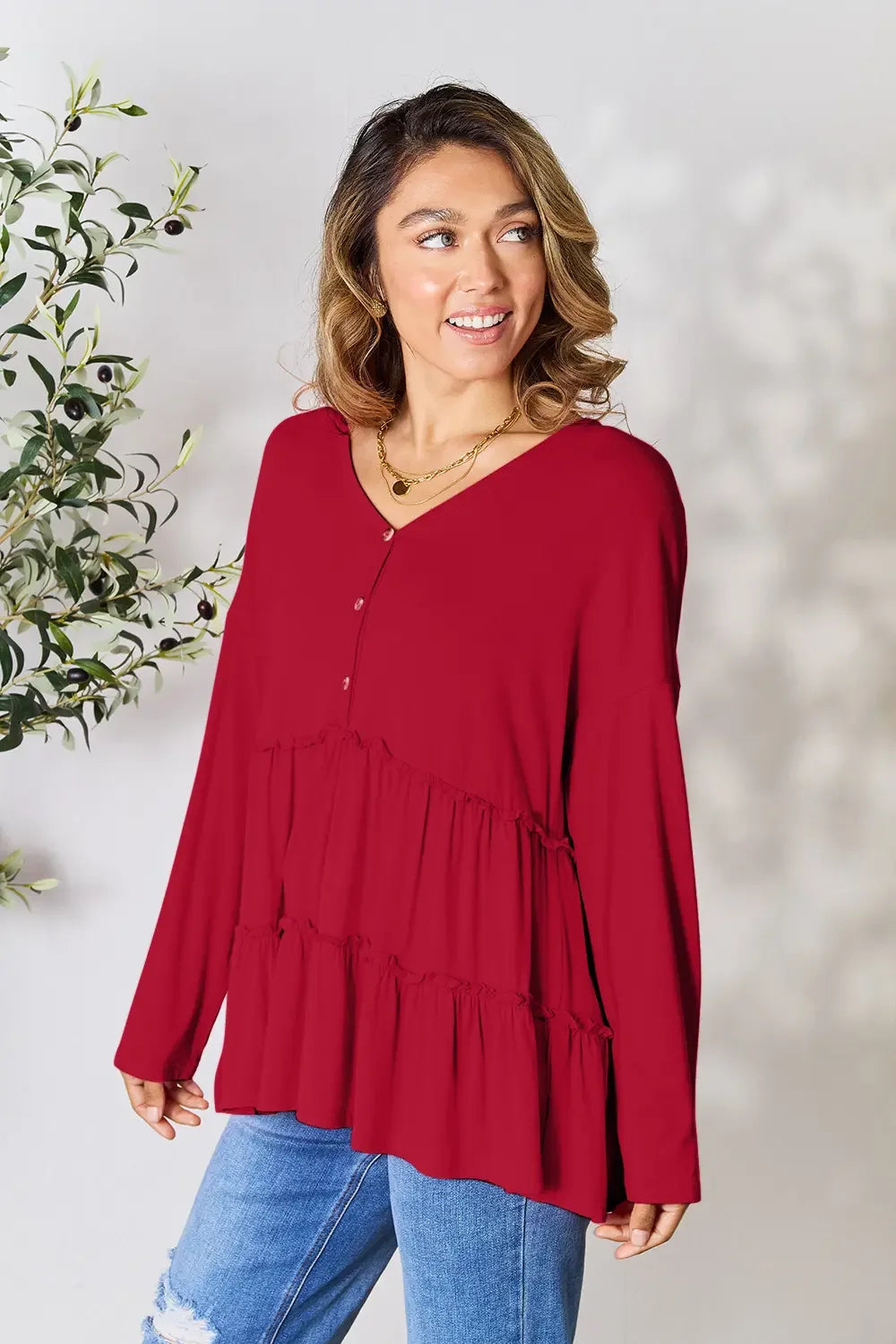Woman wearing a red long-sleeve ruffle blouse and blue distressed jeans, smiling indoors.