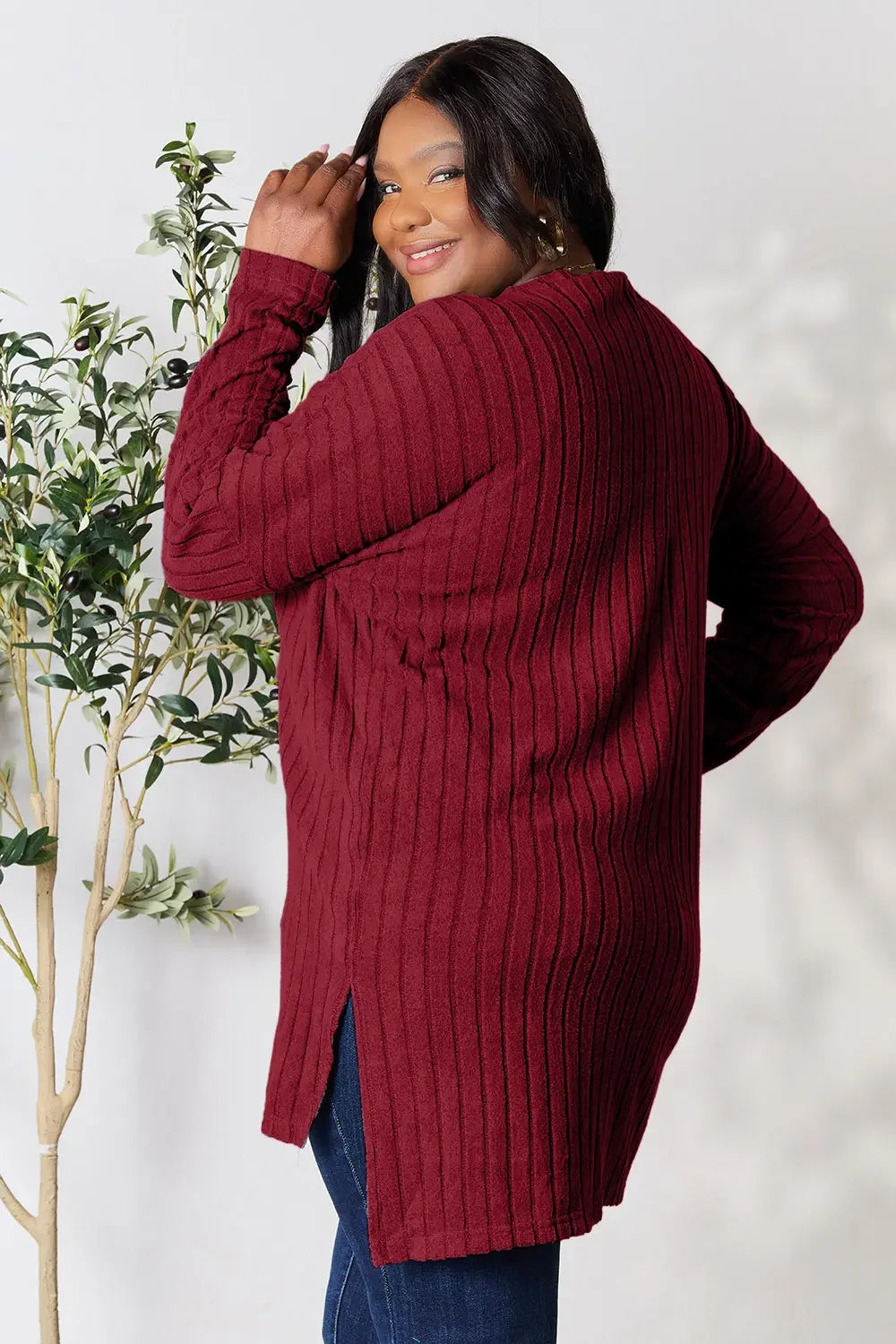 Woman in a ribbed burgundy sweater tunic and jeans, standing indoors by greenery