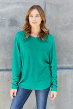 Woman wearing a green long sleeve top and blue jeans standing against a concrete wall