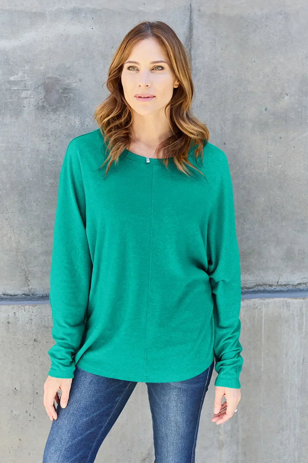 Woman wearing a green long sleeve top and blue jeans standing against a concrete wall