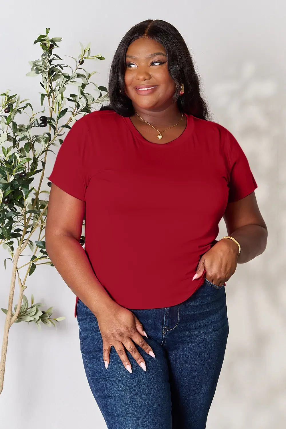 Woman in red short-sleeve top and blue jeans, smiling, standing beside a leafy plant