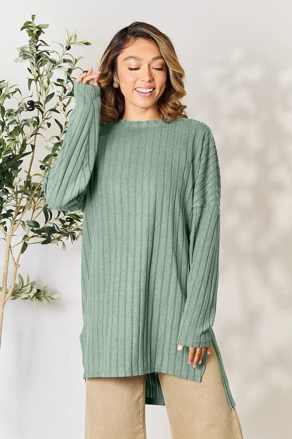 Woman wearing sage green ribbed knit sweater and beige pants, standing by an indoor plant