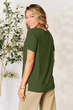 Smiling woman in olive green short sleeve top and beige pants standing by a plant