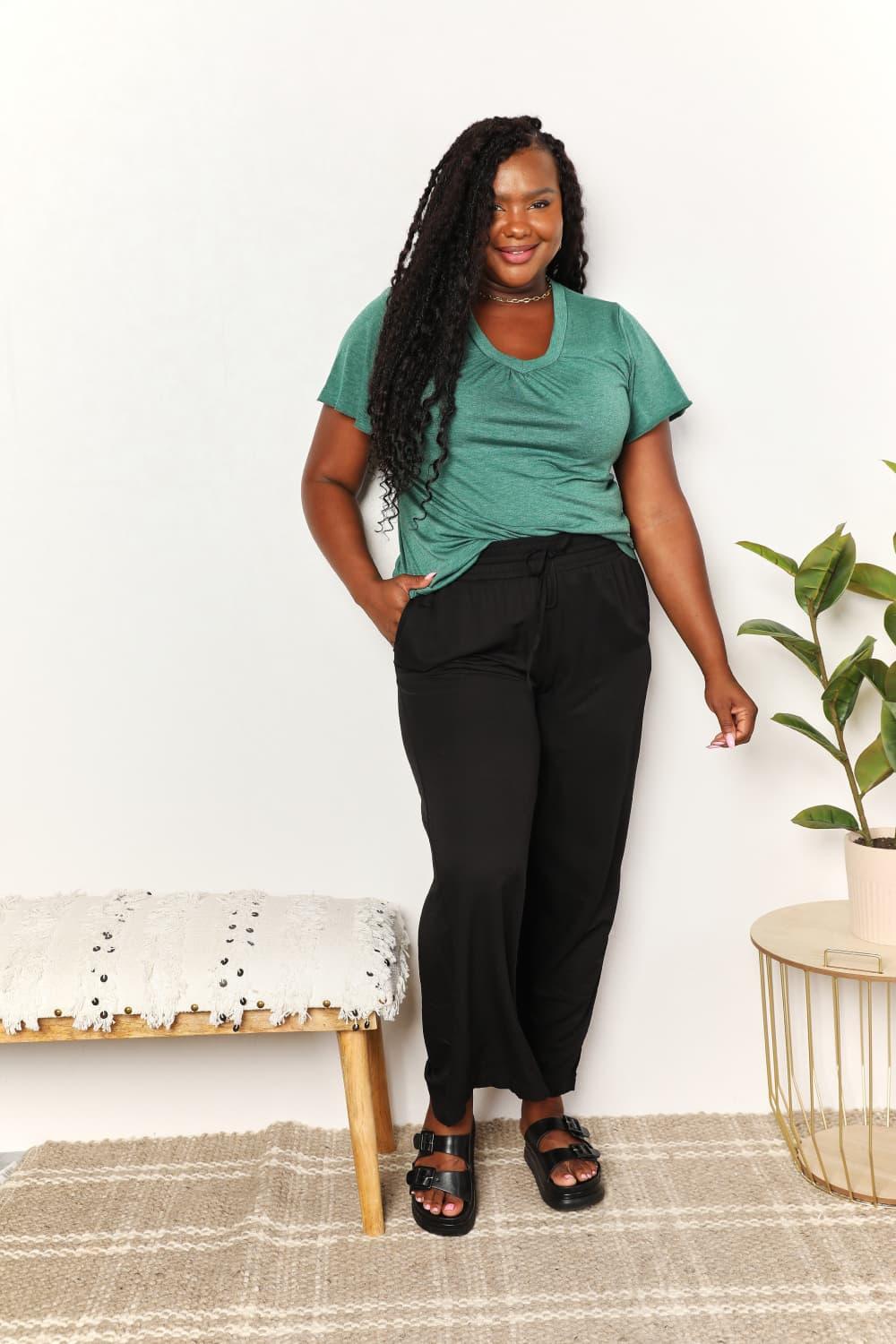 Woman in casual green t-shirt, black pants, and sandals standing indoors
