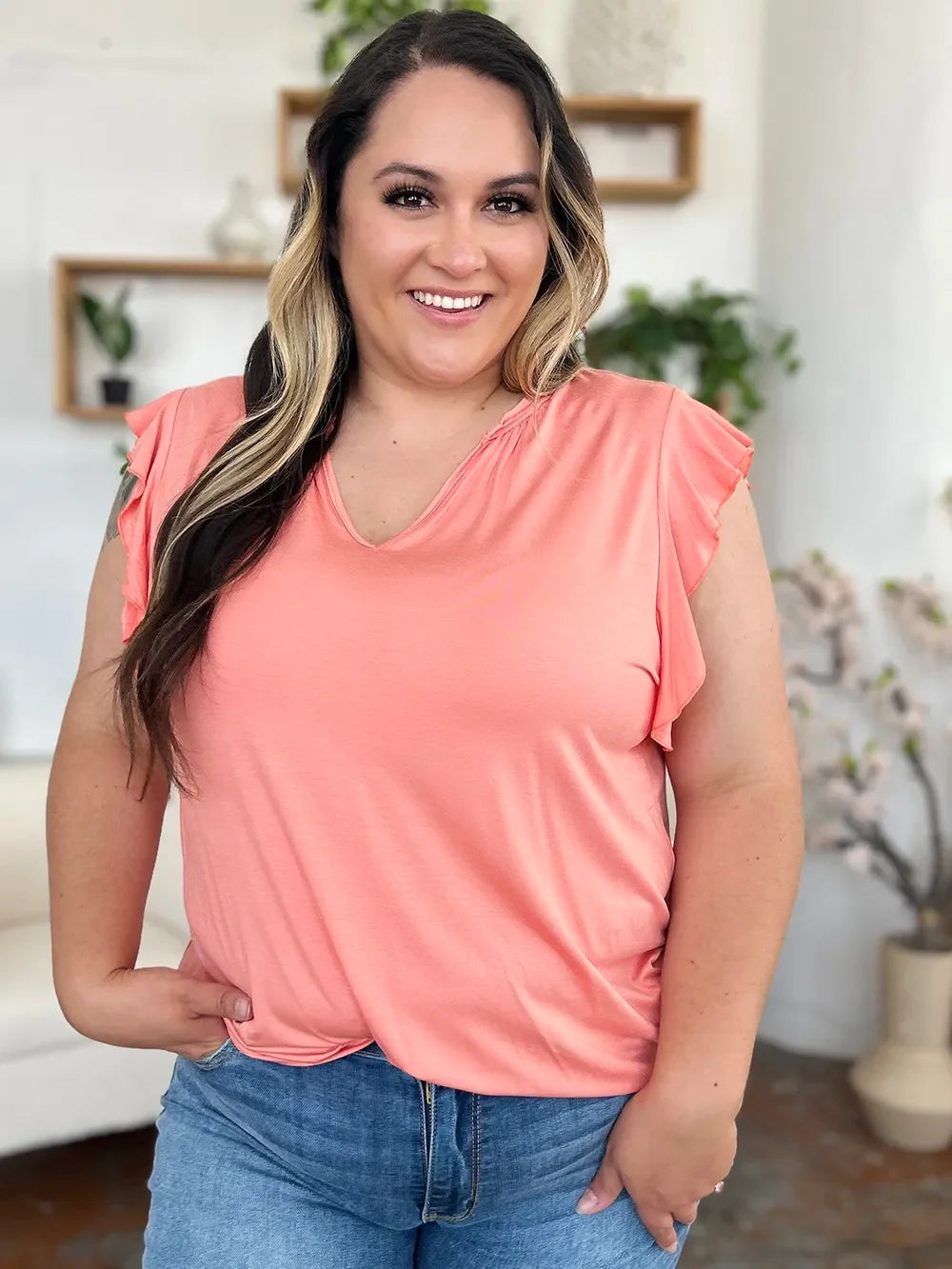 Smiling woman in a peach flutter sleeve top and blue jeans, casual indoor setting