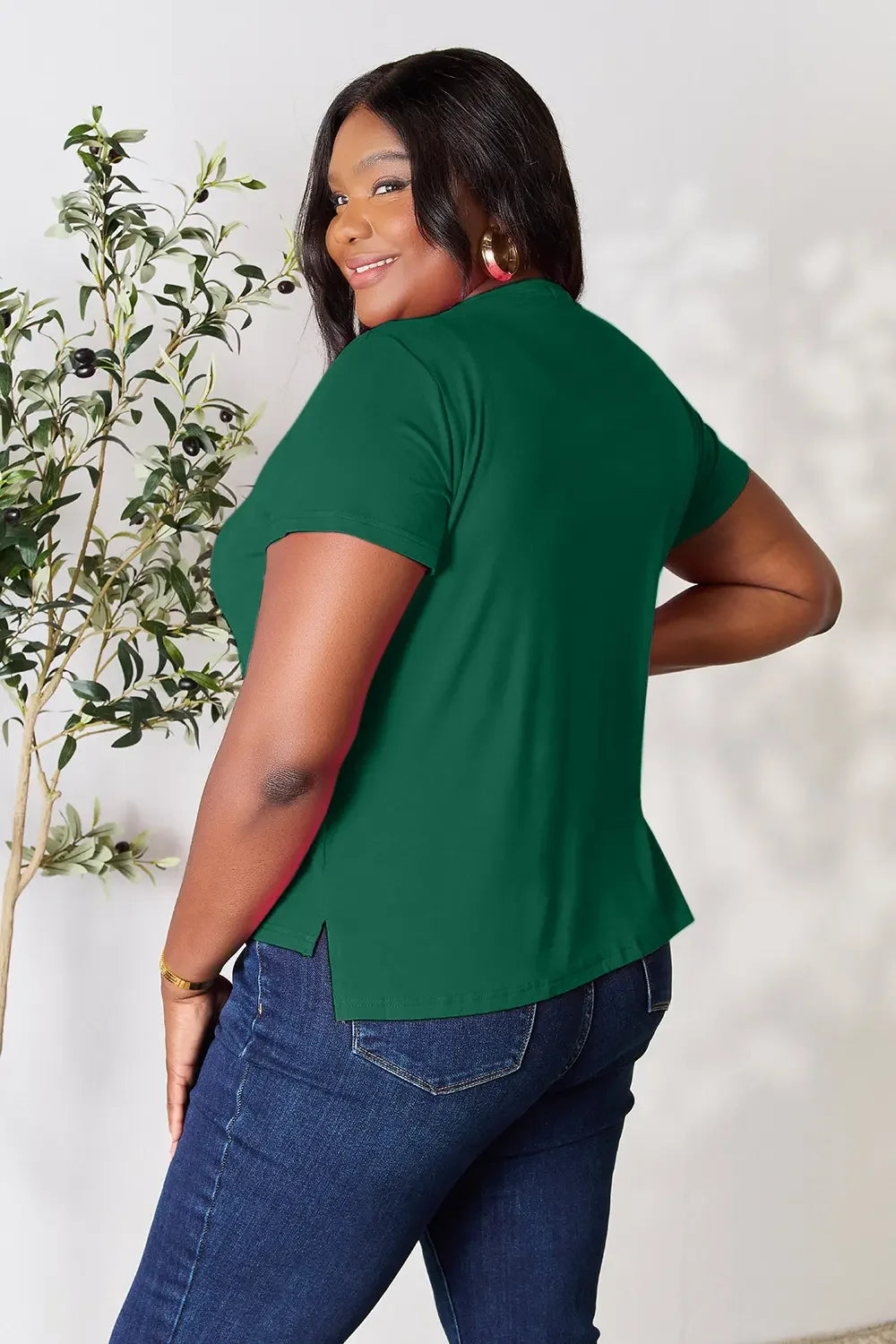 Smiling woman wearing a green short sleeve top and blue jeans, posing indoors near a plant.