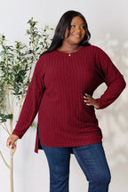 Woman wearing a burgundy ribbed sweater and blue jeans, standing by a plant