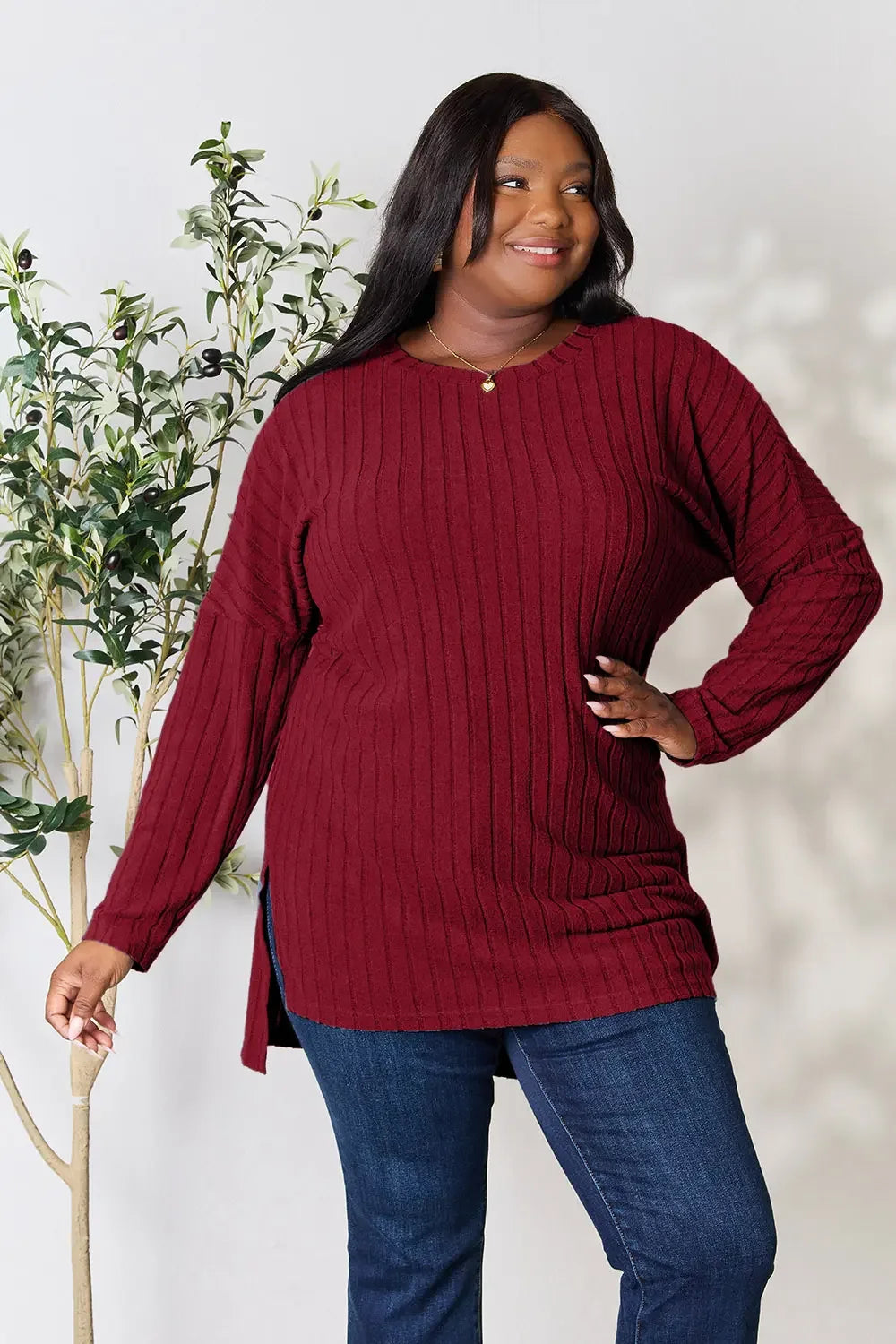Woman wearing a burgundy ribbed sweater and blue jeans, standing by a plant