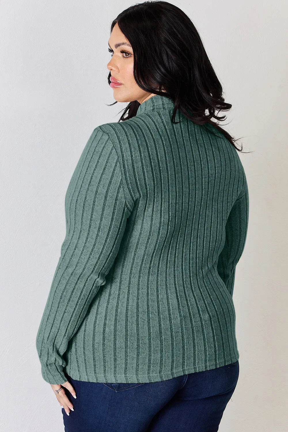 Woman wearing a green ribbed long sleeve sweater and dark blue jeans, seen from behind.