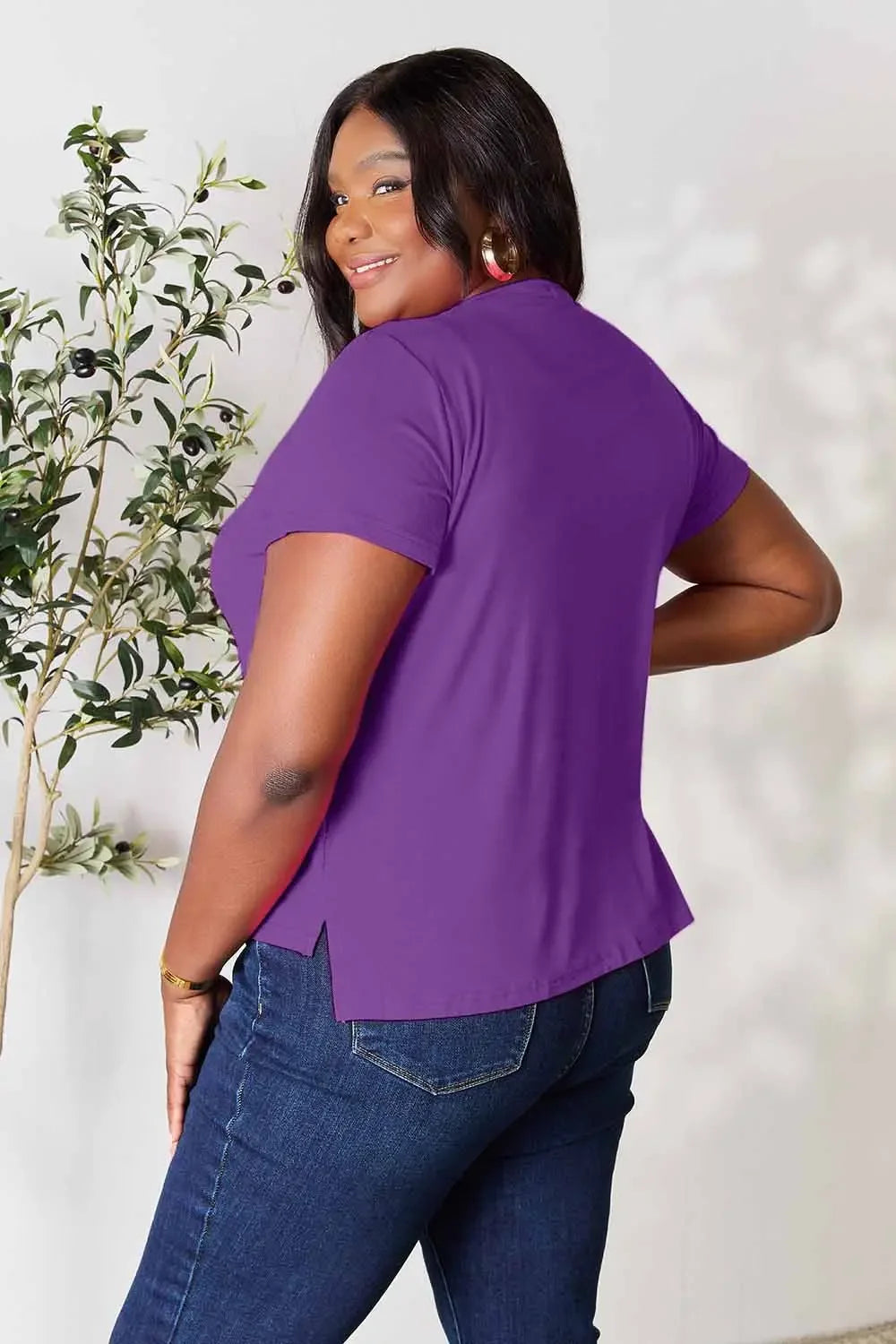 Woman in a purple short sleeve top and blue jeans, smiling, standing by a plant
