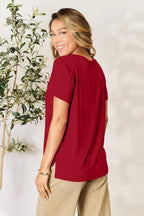 Woman wearing a red short sleeve top and beige pants, standing near a leafy plant