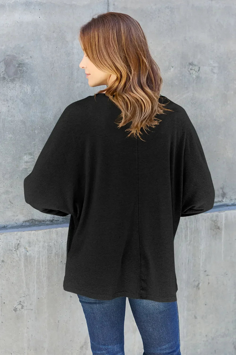 Woman in casual black long sleeve top and blue jeans standing against concrete wall
