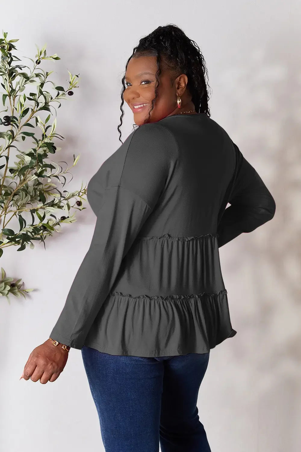 Plus size woman in long sleeve ruffle peplum top and blue jeans standing near plant