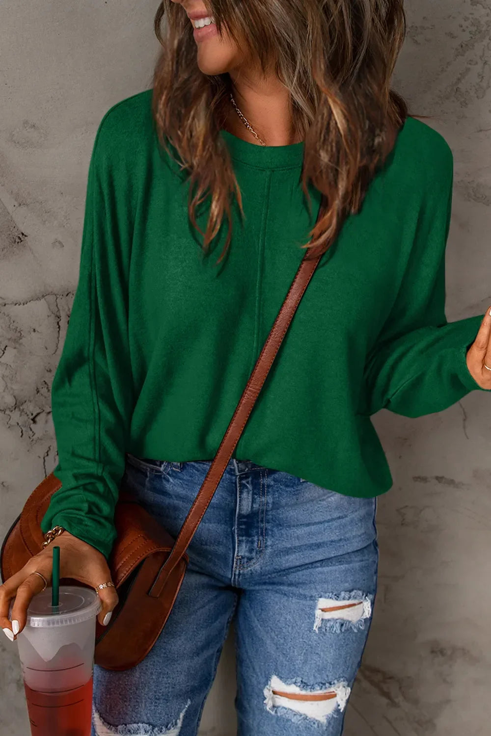 Woman in green long sleeve top and ripped blue jeans holding iced drink, crossbody bag