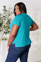 Woman in teal short sleeve top and dark blue jeans, standing indoors, smiling