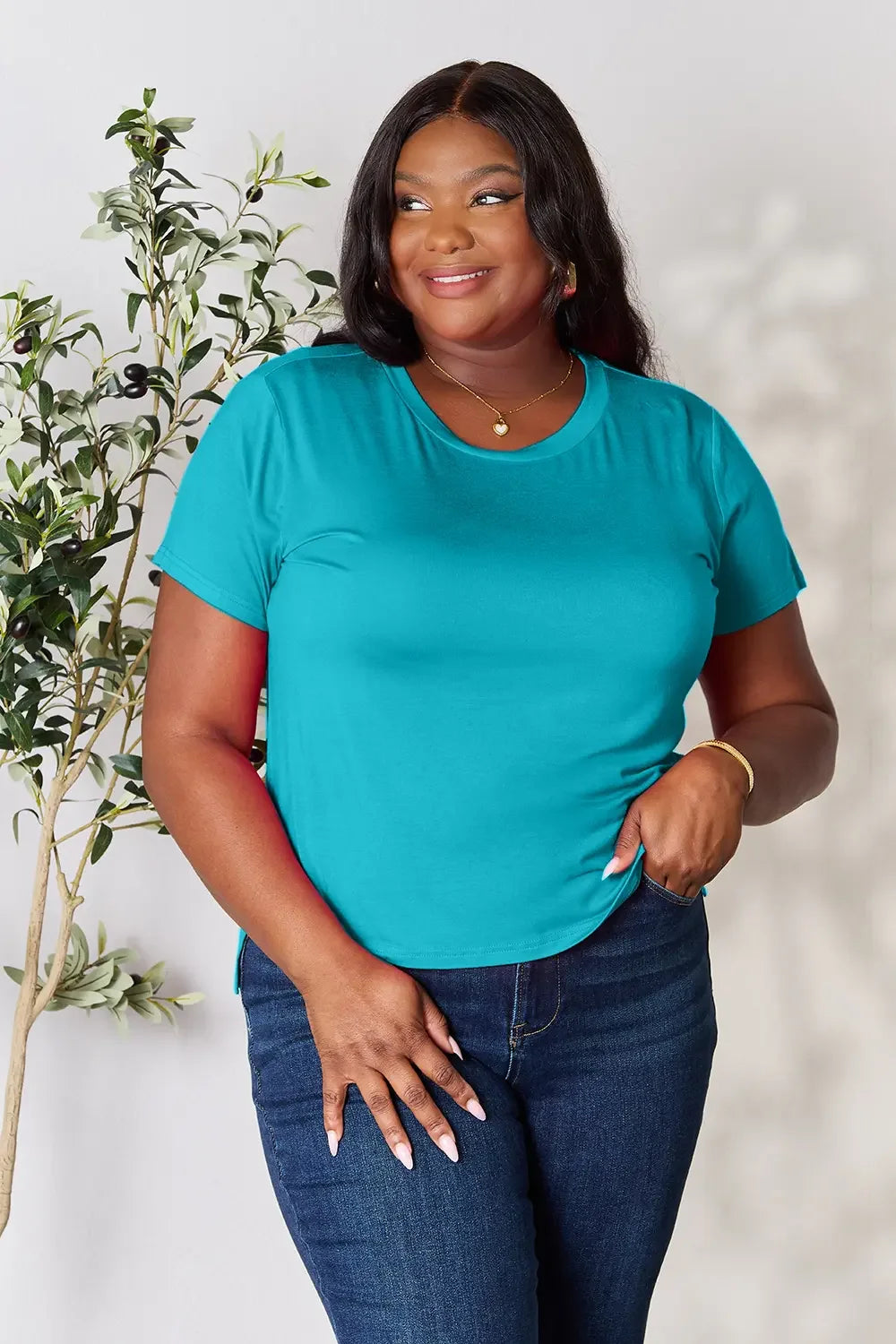 Woman wearing a teal short sleeve t-shirt and dark blue jeans, smiling indoors