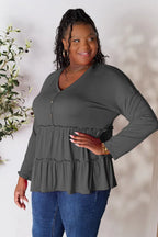 Smiling woman wearing a dark tiered long sleeve top and blue jeans, standing indoors.