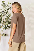 Smiling woman in a taupe short sleeve top and beige pants, standing by a leafy plant.