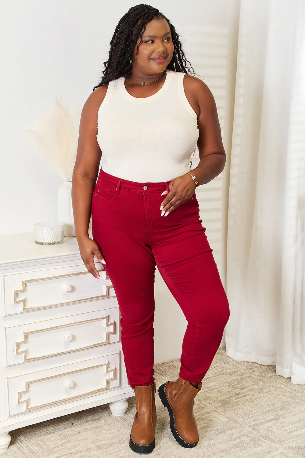 Woman in a white sleeveless top, red skinny jeans, and brown ankle boots standing indoors