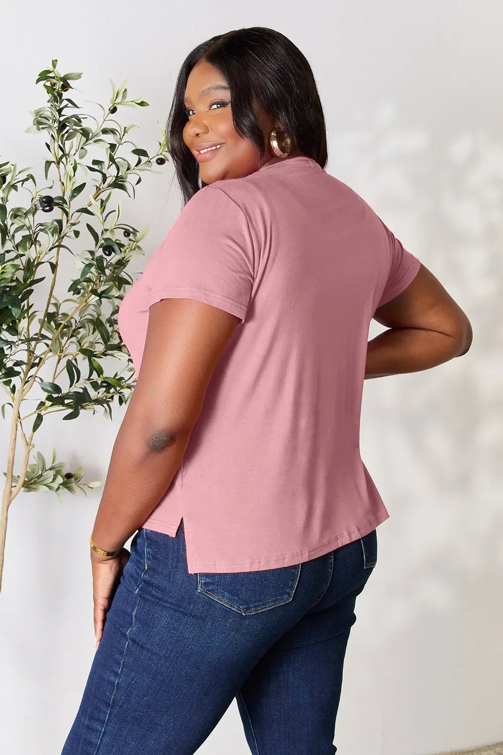 Woman in a mauve short sleeve t-shirt and blue jeans smiling, standing indoors near a plant