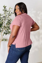 Woman in a mauve short sleeve t-shirt and blue jeans smiling, standing indoors near a plant
