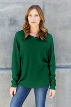 Woman wearing a green long sleeve top and blue jeans against a concrete wall