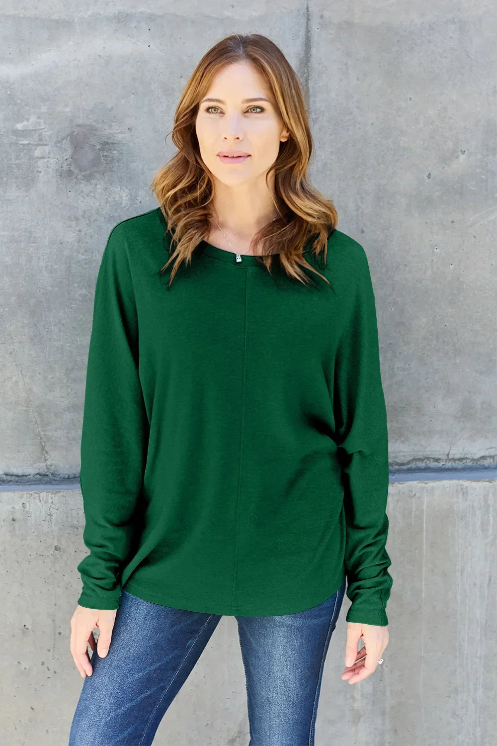 Woman wearing a green long sleeve top and blue jeans against a concrete wall