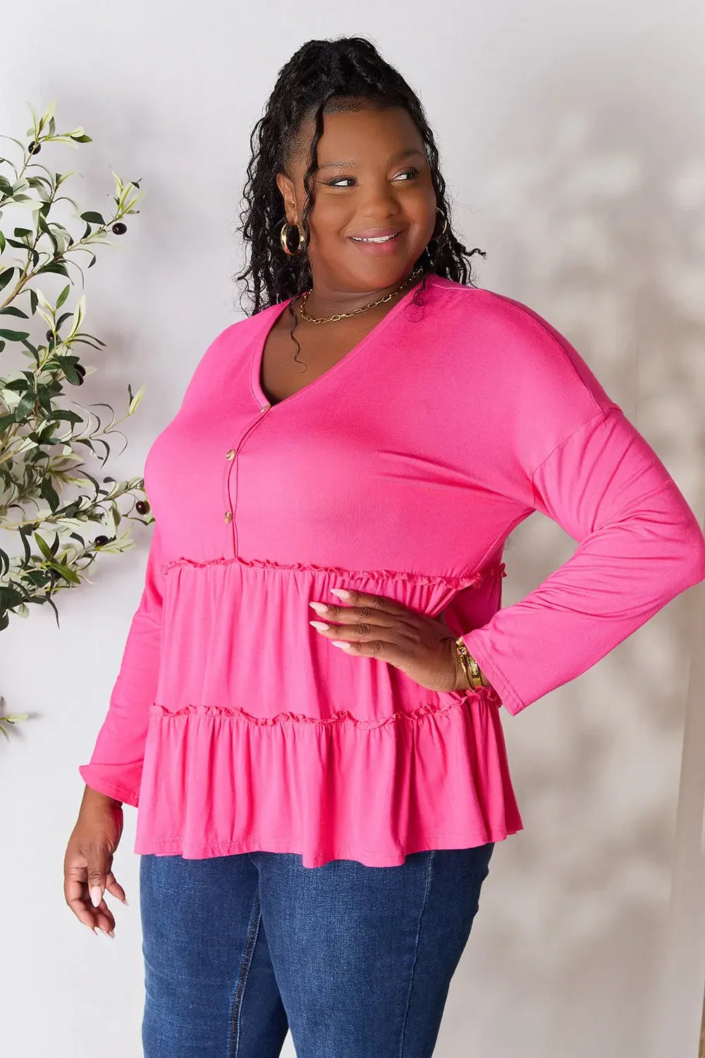 Smiling woman in long sleeve pink tiered blouse and blue jeans standing indoors