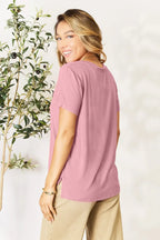 Woman wearing a mauve short sleeve t-shirt and beige pants, smiling indoors by a leafy plant.